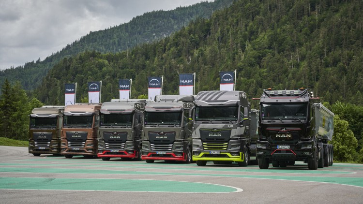 Auf einem Testgelände mit bewaldeten Bergen im Hintergrund und vor mehreren Fahnen mit MAN-Logo stehen verschiedene Lkw-Modelle des Herstellers