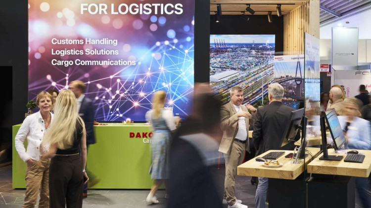 Messeatmosphäre auf der Transport Logistic 2025, Besucher gehen vor einem Stand vorbei oder stehen davor und unterhalten sich, auf dem Stand ist eine Wand groß mit einem Bild mit digitalen Netzknotenpunkten in blauen und riten Lichtern zu sehen, überschri