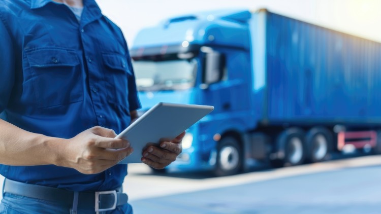Im Vordergrund hält eine Person in blauer Arbeitskleidung ein Tablet. Im Hintergrund steht ein blauer Sattelzug mit Kofferauflieger auf einer befestigten Fläche.