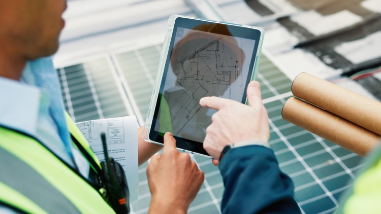 Solarmodul auf einem Dach, unscharf im Hintergrund, davor stehen zwei Personen in gelben Warnwesten und zeigen auf ein Tablet. Die eine hält braune Papierrollen mit Plänen in der anderen Hand