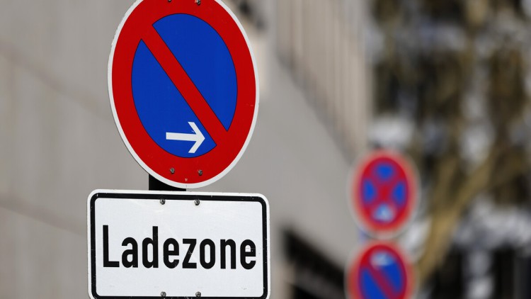 Ladezonenschild im Bildvordergrund, Bildhintergrund unscharf städtische Gebäude an einer Straße. Eine Ladezone ist ein speziell gekennzeichneter Bereich im Straßenverkehr (oft mit Halteverbotsschildern und Zusatztafeln); der primär dem schnellen Be- und E