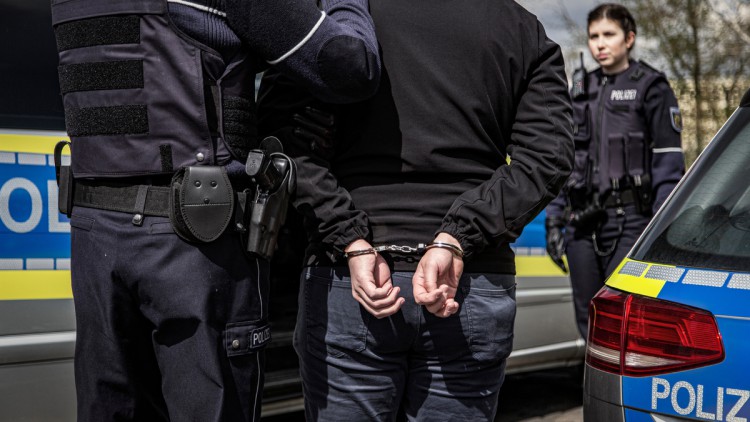Zwei Polizisten verhaften einen Kriminellen. Im Bildvordergrund stehen zwischen zwei Polizeifahrzeugen ein Polizist und der Verbrecher mit Handschellen mit dem Rücken zum Betrachter. Im Hintergrund steht eine Polizistin und blickt ihrem Kollegen und dem V