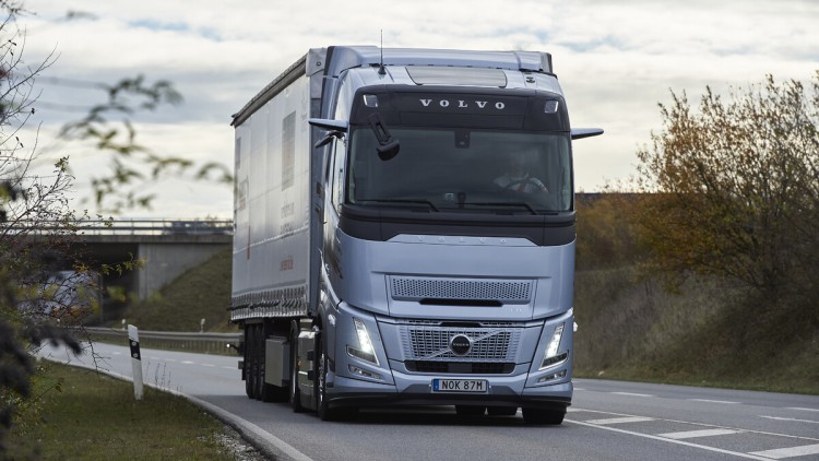 Ein Volvo FH Aero I-Save fährt bei bedecktem Himmel und schummerigen Lichtverhältnissen auf einer Straße durch die Landschaft, von vorne aufgenommen, zu sehen ist eine buschbestandene Böschung und im Hintergrund eine Brücke