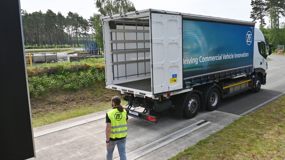 Ein Lkw fährt mit geöffneten Hecktüren des Laderaums rückwärts an eine Rampe. Eine Person mit Warnweste kreuzt den Weg des Lkw